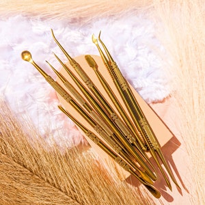 May include: A set of eight gold metal tools with different shaped tips, arranged on a light brown surface. The tools are used for crafting and art projects.