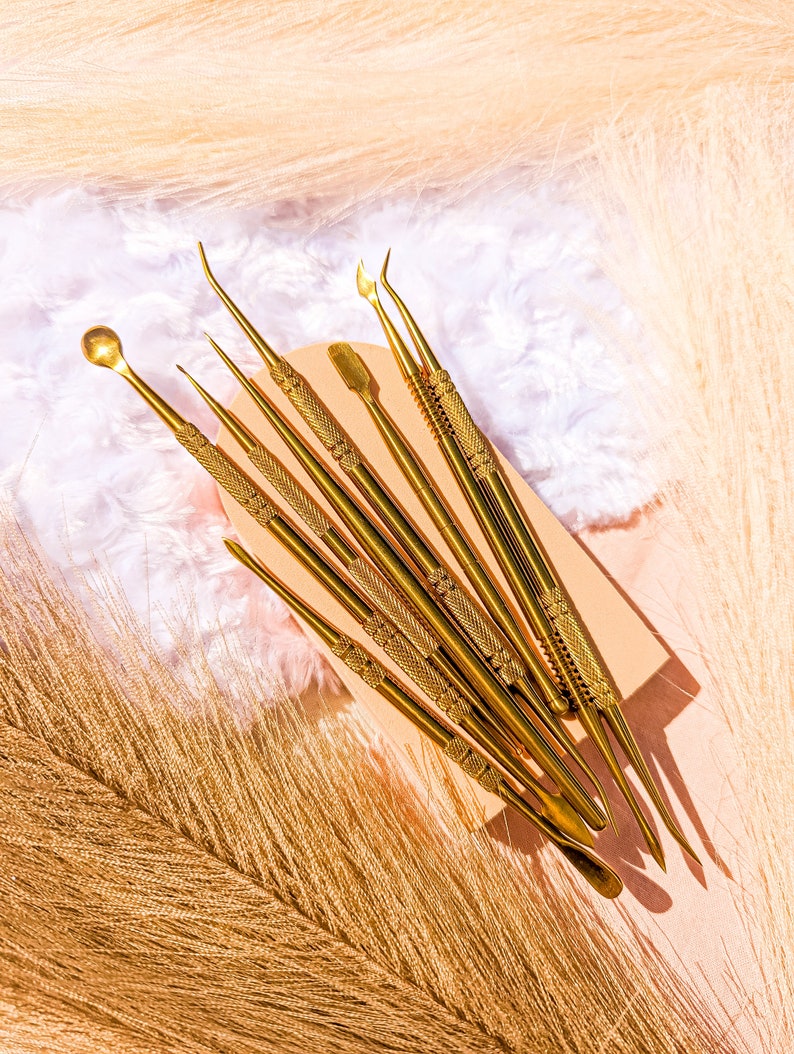 May include: A set of eight gold metal tools with different shaped tips, arranged on a light brown surface. The tools are used for crafting and art projects.