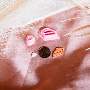 Puede incluir: Un conjunto de cinco cortadores de galletas de pl&aacute;stico rosa y naranja con forma de coraz&oacute;n, una rebanada de pastel, una tarta y un coraz&oacute;n peque&ntilde;o. Los cortadores est&aacute;n sobre un fondo rosa y blanco.