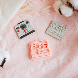 May include: Three vintage-style camera shaped cookie cutters, one pink, one gray, and one light gray. The cutters are on a pink fabric background with white cotton balls.