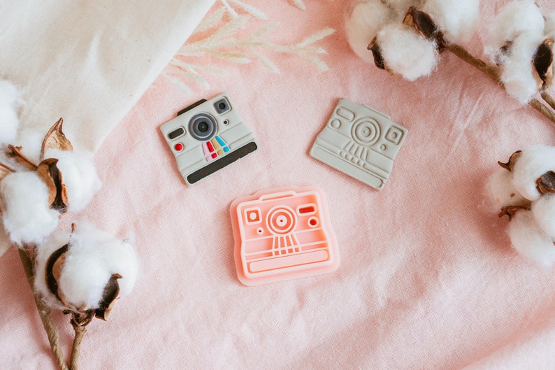 May include: Three vintage-style camera shaped cookie cutters, one pink, one gray, and one light gray. The cutters are on a pink fabric background with white cotton balls.