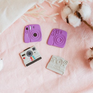 May include: Four miniature camera shaped toys on a pink fabric background. Two of the cameras are purple and two are gray.