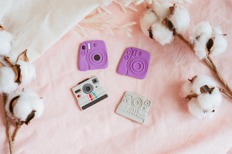 May include: Four miniature camera shaped toys on a pink fabric background. Two of the cameras are purple and two are gray.