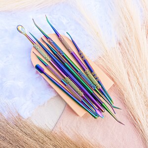May include: A set of nine iridescent metal tools with different shaped tips, arranged on a wooden block. The tools are used for nail art and cuticle care.
