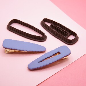 Op de afbeelding: Vier haarclips, twee zijn paars en twee zijn bruin. De paarse clips hebben een getextureerd oppervlak en een gouden metalen sluiting. De bruine clips zijn glad en hebben een afgeronde vorm.