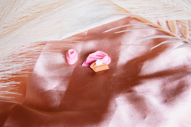 Puede incluir: Dos dijes en forma de pastel rosa y blanco y un dije en forma de pastel marr&oacute;n y blanco sobre un fondo de tela color melocot&oacute;n.