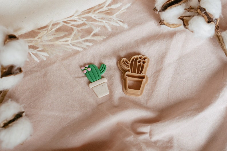Peut inclure: Deux emportes-pi&egrave;ces en forme de cactus, l'un vert et l'autre brun, sur un fond de tissu rose clair. L'emporte-pi&egrave;ce vert a une fleur rose sur le dessus et est dans un pot blanc. L'emporte-pi&egrave;ce brun est une repr&eacute;sentation plus abstraite d'un cactus.
