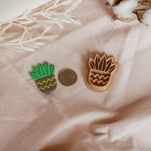 May include: Two brown plastic cookie cutters shaped like a potted succulent plant. The cutters are on a light pink fabric surface.