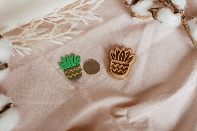 May include: Two brown plastic cookie cutters shaped like a potted succulent plant. The cutters are on a light pink fabric surface.
