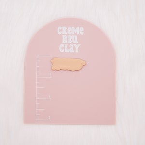 May include: A pink, semi-circular, plastic crafting mat with a ruler on the side. The mat has the text "CREME BRU CLAY" printed on it. A small, light brown, clay cutout of the island of Puerto Rico is on the mat.