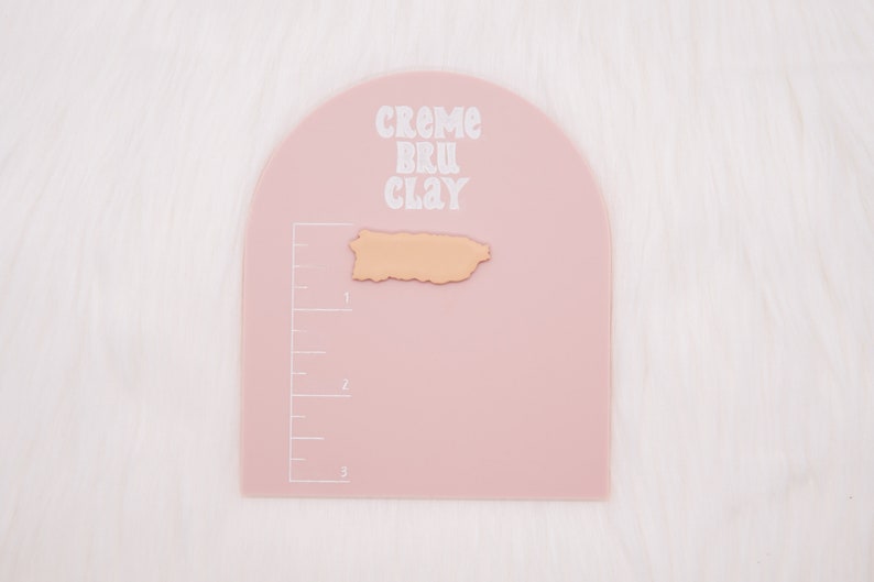 May include: A pink, semi-circular, plastic crafting mat with a ruler on the side. The mat has the text "CREME BRU CLAY" printed on it. A small, light brown, clay cutout of the island of Puerto Rico is on the mat.