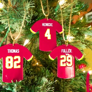 Puede incluir: Tres adornos navideños rojos con forma de camiseta y detalles amarillos cuelgan de una cuerda en un árbol de Navidad. Cada adorno muestra un nombre y un número diferentes: Heinicke #4, Thomas #82 y Fuller #29. Festivos y decorativos.