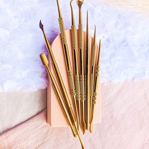 May include: A set of eight gold-toned metal tools with various shapes and sizes. The tools are arranged on a light pink surface.