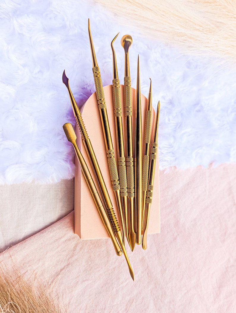 May include: A set of eight gold-toned metal tools with various shapes and sizes. The tools are arranged on a light pink surface.