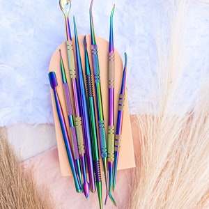 May include: A set of nine iridescent metal tools with various shapes and sizes, arranged on a light brown surface. The tools are used for crafting and nail art.