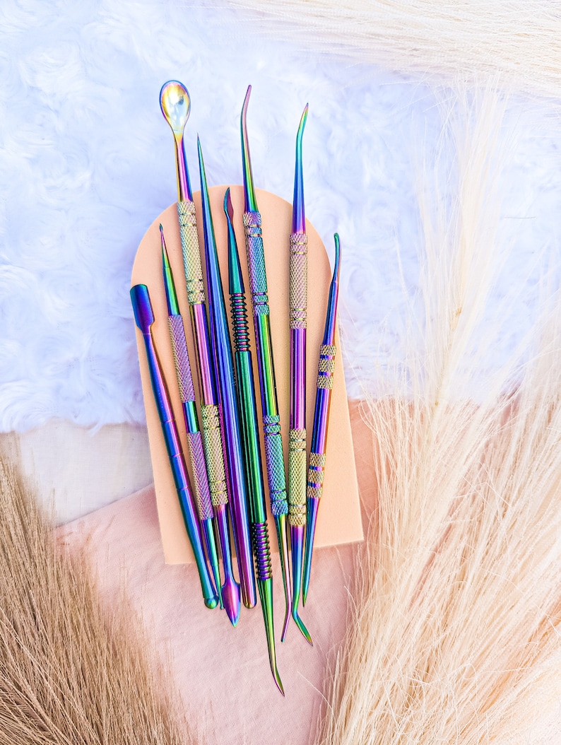 May include: A set of nine iridescent metal tools with various shapes and sizes, arranged on a light brown surface. The tools are used for crafting and nail art.