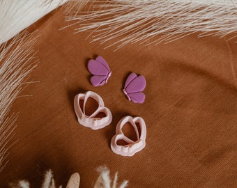 May include: Two purple butterfly-shaped cookie cutters and two pink flower-shaped cookie cutters on a brown fabric background.