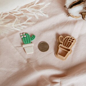Peut inclure: Deux emportes-pi&egrave;ces en forme de cactus, l'un vert et l'autre brun, sur un fond de tissu rose. Le cactus vert a une fleur rose sur le dessus.
