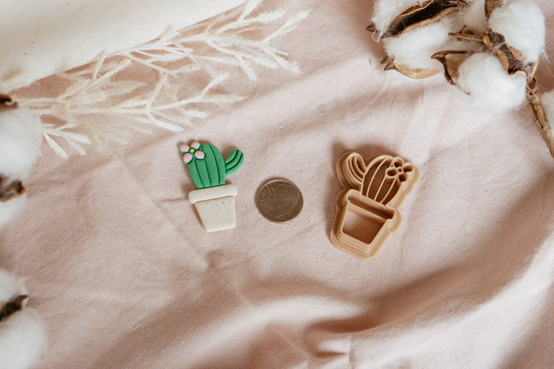 Peut inclure: Deux emportes-pi&egrave;ces en forme de cactus, l'un vert et l'autre brun, sur un fond de tissu rose. Le cactus vert a une fleur rose sur le dessus.