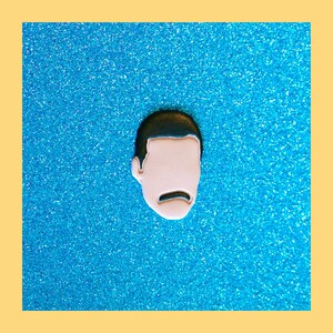 May include: A black and white enamel pin featuring a cartoon face with a mustache. The pin is on a blue sparkly background.