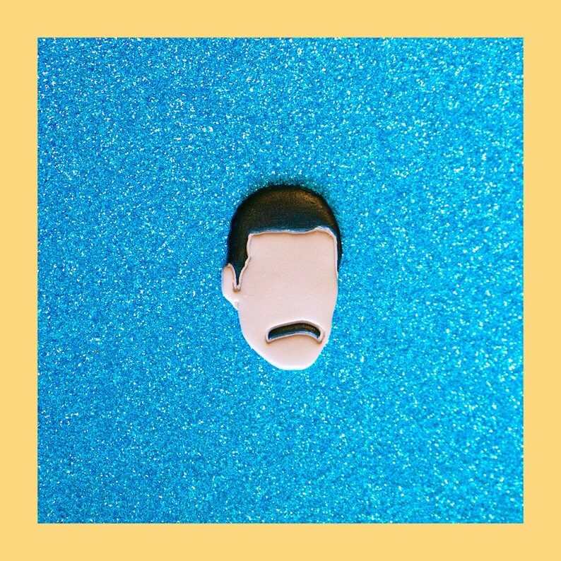 May include: A black and white enamel pin featuring a cartoon face with a mustache. The pin is on a blue sparkly background.