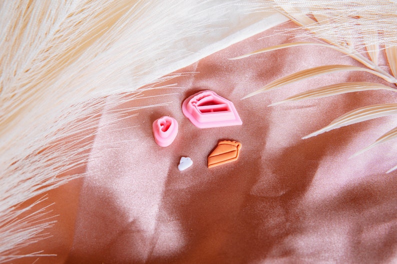 Puede incluir: Cuatro cortadores de galletas de pl&aacute;stico rosa y naranja con forma de coraz&oacute;n, una rebanada de pastel, una rebanada de pastel y una nube. Los cortadores est&aacute;n sobre un fondo de tela marr&oacute;n claro.