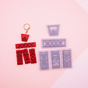 May include: A set of red and clear plastic molds for making polymer clay earrings. The molds feature geometric designs and are arranged on a pink surface.