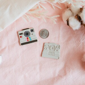 May include: Two white plastic toy cameras on a pink surface with cotton bolls. One camera has a rainbow stripe design and the other is plain.