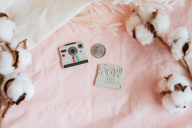 May include: Two white plastic toy cameras on a pink surface with cotton bolls. One camera has a rainbow stripe design and the other is plain.