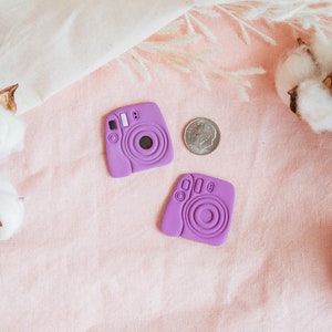 May include: Two purple miniature instant camera charms on a pink fabric background with cotton bolls.