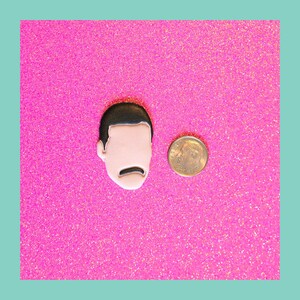 May include: A black and white enamel pin featuring a cartoon portrait of a man with a mustache. The pin is on a pink glitter background.