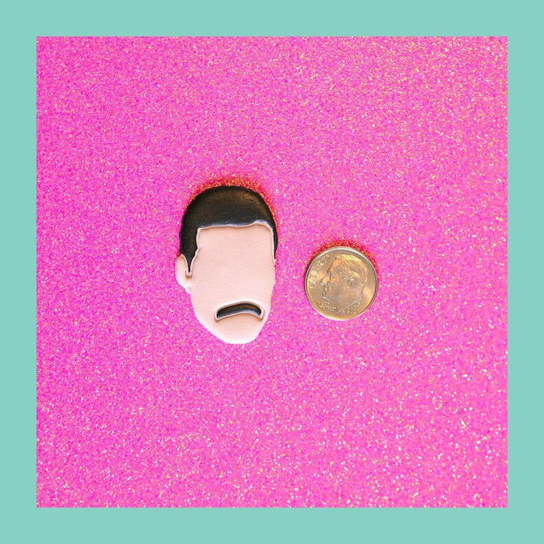 May include: A black and white enamel pin featuring a cartoon portrait of a man with a mustache. The pin is on a pink glitter background.