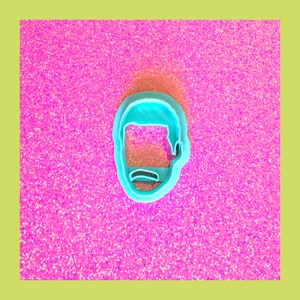 May include: A blue plastic cookie cutter shaped like a person's head with a simple design. The cutter is on a pink sparkly background.