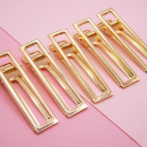 Peut inclure: Six pinces à cheveux en métal doré de forme rectangulaire avec un mécanisme à ressort. Les pinces sont disposées en rangée sur un fond rose.