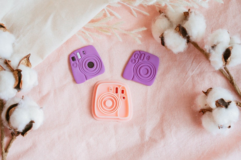 May include: Two purple and one pink plastic camera shaped molds on a pink fabric background with cotton boll stems.