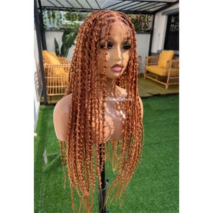 Puede incluir: Peluca trenzada marrón exhibida en una cabeza de maniquí. La peluca presenta largas trenzas individuales con una línea de cabello de aspecto natural. El fondo incluye muebles de exterior y vegetación.