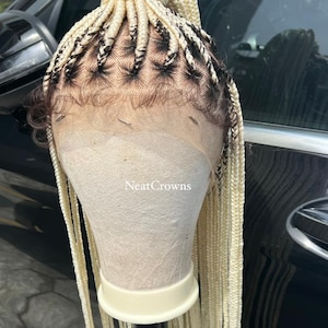 Puede incluir: Una peluca trenzada rubia con un frente de encaje y una coleta. La peluca está en una cabeza de maniquí y está peinada en un patrón de trenza recta y ordenada. La peluca está etiquetada como "NeatCrowns".