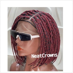Peut inclure: Une perruque tressée de couleur bordeaux et noire avec une dentelle frontale. La perruque est coiffée en un bob court avec des tresses se terminant par des bords nets. La perruque est sur une tête de mannequin et le texte "NeatCrowns" est visible en bas à droite de l'image.