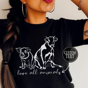 Puede incluir: Camiseta negra con una silueta blanca de una vaca y un cerdo, y el texto "love all animals" con una flecha que apunta a los animales. El texto "CUSTOM TEXT" está en un círculo.