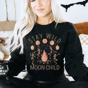 K&ouml;nnte beinhalten: Ein schwarzes Sweatshirt mit dem Text "Stay Wild Moon Child" in goldenen Buchstaben. Das Design zeigt ein Lagerfeuer mit Skeletten, die darum herum tanzen, und einen Halbmond mit Sternen.