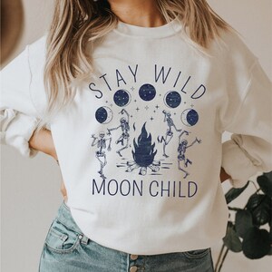K&ouml;nnte beinhalten: Wei&szlig;er Pullover mit einem blauen Grafikdruck von Skeletten, die um ein Lagerfeuer tanzen, mit dem Text "Stay Wild Moon Child".