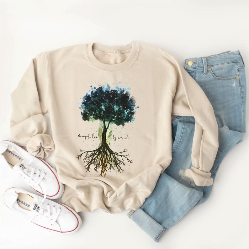 Puede incluir: Una sudadera beige con un dise&ntilde;o de &aacute;rbol de acuarela. El &aacute;rbol tiene hojas azules, verdes y amarillas. Las ra&iacute;ces del &aacute;rbol son marrones y se extienden. Las palabras "biophilia" y "spirit" est&aacute;n escritas a ambos lados del &aacute;rbol.