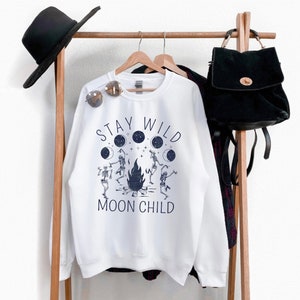K&ouml;nnte beinhalten: Wei&szlig;er Pullover mit einem grafischen Aufdruck von Skeletten, die um ein Lagerfeuer tanzen, mit dem Text "Stay Wild Moon Child".