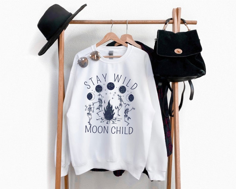 K&ouml;nnte beinhalten: Wei&szlig;er Pullover mit einem grafischen Aufdruck von Skeletten, die um ein Lagerfeuer tanzen, mit dem Text "Stay Wild Moon Child".