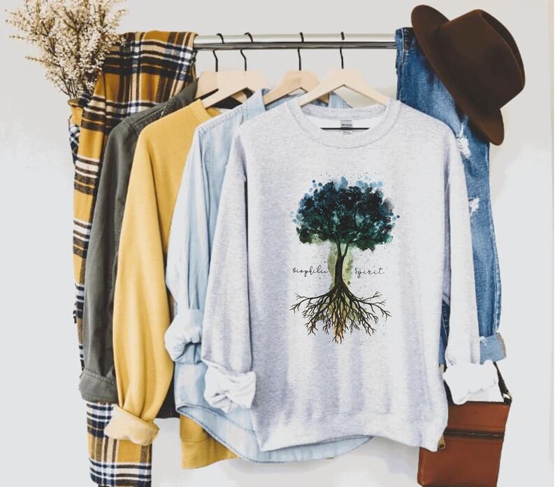 Puede incluir: Una sudadera gris con un dise&ntilde;o de &aacute;rbol de acuarela y el texto "biophilia spirit" impreso en la parte delantera.