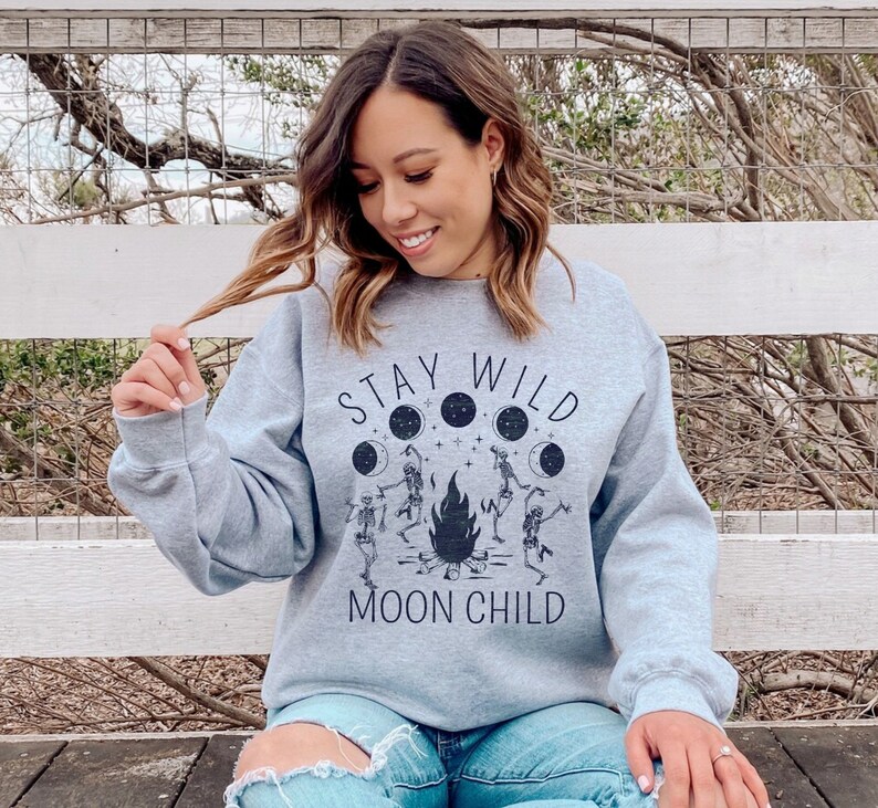 K&ouml;nnte beinhalten: Ein graues Sweatshirt mit dem Text "Stay Wild Moon Child" und einer Grafik von Skeletten, die um ein Feuer mit Monden im Hintergrund tanzen.
