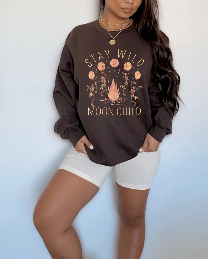 K&ouml;nnte beinhalten: Ein braunes Sweatshirt mit einem grafischen Aufdruck von Skeletten, die um ein Lagerfeuer tanzen, mit dem Text "Stay Wild Moon Child".