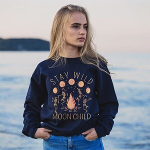 K&ouml;nnte beinhalten: Ein marineblauer Pullover mit einem grafischen Aufdruck von Skeletten, die um ein Lagerfeuer tanzen, mit dem Text "Stay Wild Moon Child".