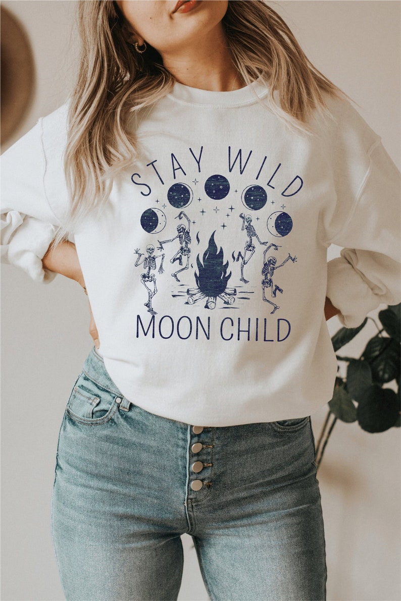 K&ouml;nnte beinhalten: Wei&szlig;er Pullover mit einem blauen Grafikdruck von Skeletten, die um ein Lagerfeuer tanzen, mit dem Text "Stay Wild Moon Child".