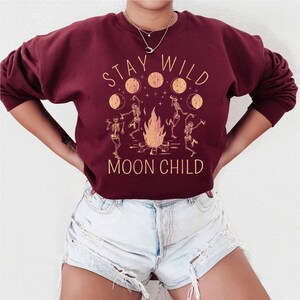K&ouml;nnte beinhalten: Ein weinroter Pullover mit dem Text "Stay Wild Moon Child" in gelber Schrift. Die Grafik zeigt ein Lagerfeuer mit tanzenden Skeletten und Monden.